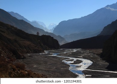 A glittering, winding river flows calmly through a great canyon along the Annapurna Range in the himalyas near Manang, Nepal in Asia. Great mountains appear in shades of blue towards the horizon.