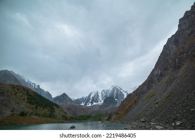 Gloomy Autumn Landscape With Mountain Lake Against Large Snowy Mountains Under Gray Cloudy Sky. Dark Atmospheric Scenery With Alpine Lake And Snow Mountain Range And Peaked Top In Overcast Weather.