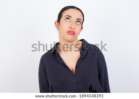 Gloomy, bored young beautiful Arab woman wearing gray dress against white studio background frowns face looking up, being upset with so much talking hands down, feels tired and wants to leave.