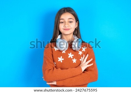 Gloomy dissatisfied beautiful kid girl wearing knitted orange sweater over blue background looks with miserable expression at camera from under forehead, makes unhappy grimace
