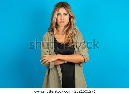 Gloomy dissatisfied young beautiful blonde woman wearing overshirt looks with miserable expression at camera from under forehead, makes unhappy grimace
