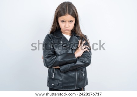 Gloomy dissatisfied Young beautiful teen girl wearing biker jacket looks with miserable expression at camera from under forehead, makes unhappy grimace