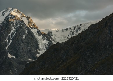 Gloomy Landscape With Giant Snowy Mountain Range And Sunlit Gold Rocks In Dramatic Gray Sky. Huge Snow Mountains With Hanging Glacier And Cornice Under Rainy Clouds. Snow-covered Mountains In Overcast