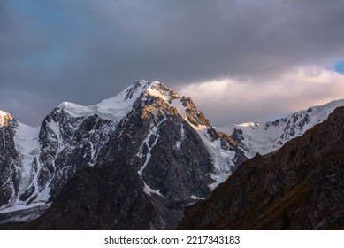 Gloomy Landscape With Giant Snowy Mountain Top And Sunlit Gold Rocks In Dramatic Gray Sky. Huge Snow Mountains With Hanging Glacier And Cornice Under Rainy Clouds. Snow-covered Mountains In Overcast.