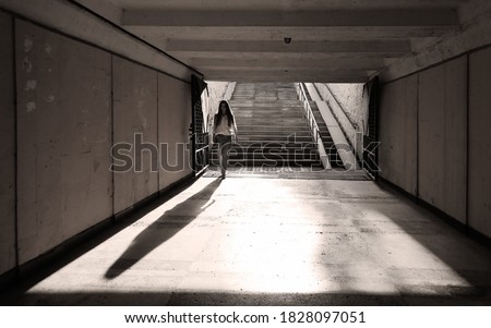 Gloomy long pedestrian underpass tunnel in dark colors. Young woman, illuminated by backlight, went down and walks alone, not expecting danger