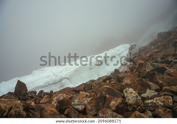 Gloomy minimalist foggy mountain landscape with snow\
cornice over abyss inside cloud. Above snow cornice on abyss edge\
in dense fog. Dangerous mountain pass above precipice. Danger rocks\
in thick fog.