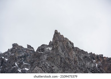 Gloomy mountain landscape with high sharp rockies with snow in gray rainy sky. Closeup of sharp rocks and peaked top in gray cloudy sky. Bleak overcast scenery with mountain range with pointy peak.