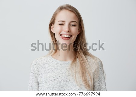 Gloomy young blonde woman with dyed hair dressed casually making faces at camera, blinking, sticking out her tongue. Positive girl having fun indoors