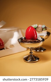A glossy red Easter egg in a silver cup, surrounded by chocolate strands and a delicate flower.