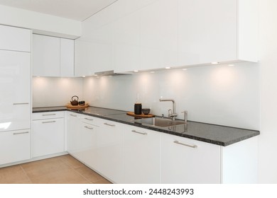 Glossy white minimalist modern kitchen with a black marble countertop. Spotlights illuminate the countertop, above which are two trays, one with a teapot and one with a vase. No one inside - Powered by Shutterstock