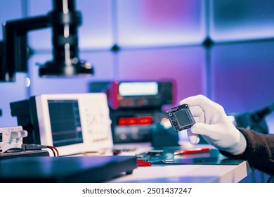 Gloved hand holding a microchip in a high-tech laboratory setting with various electronic instruments in the background.