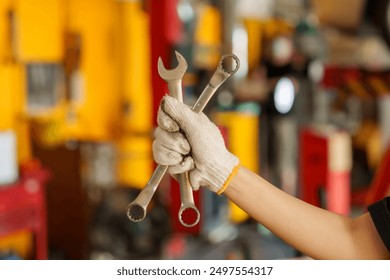 Gloved hand holding two wrenches in front of blurry automotive workshop background. Focus on tools, representing readiness and precision in mechanic work. Captures concept of expertise, preparedness, - Powered by Shutterstock