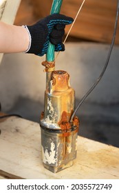 Gloved Hand Holds An Old Rusty Well Pump With A Hose In Draw Well. Pump Repair