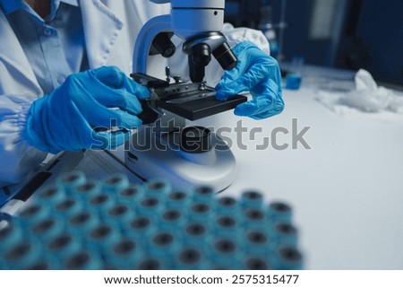 Gloved hands adjusting a microscope slide on a microscope stage, with a blurred rack of test tubes in the foreground, create a scene of focused scientific investigation