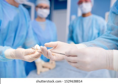 Gloved hands of assistant passing sterile steel instrument to surgeon during operation in surgery room - Powered by Shutterstock