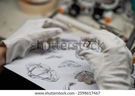 Gloved hands holding gemstone while sketching various ring designs on paper with gemstones and tools on workbench creating intricate jewelry pieces