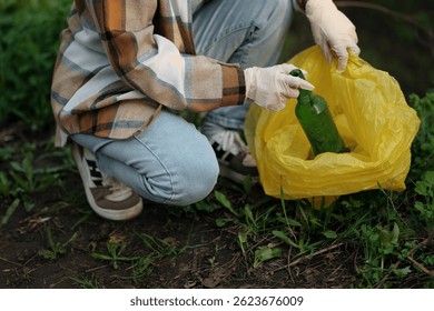 Un hombre con guantes se pone en cuclillas y recoge la basura en un bosque o parque, colocando una botella de vidrio en una bolsa de basura amarilla en Clima cálido. Concepto de limpieza ambiental, reciclaje y voluntariado