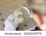 Gloved specialist examining gold coin with magnifying glass. Numismatist looking at coin trough magnifying glass for valuing coin.