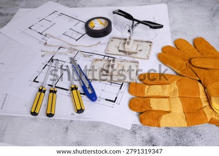 Gloves, screwdrivers, cutter and electrical components arranged over blueprints, illustrating installation, repair and maintenance processes in construction.
