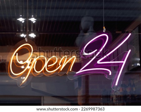 Glowing ad is OPEN 24 hours a day in cafeteria window. Modern electric signboard to attract attention of passers-by. Dark interior and colorful illuminated sign.