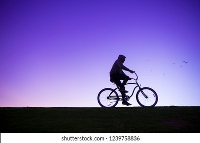 Glowing Backlit Silhouette Of Man In Hoodie Cycling On A Beach Cruiser Bicycle Against Purple Sunset Sky