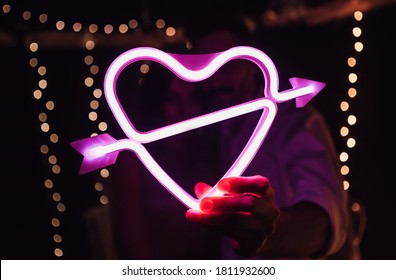 Glowing bright luminous neon heart sign pierced by cupid arrow, amour shape in vivid colors. Newlyweds holding digital shiny love target. Lovely young couple have a date, spending time together. - Powered by Shutterstock