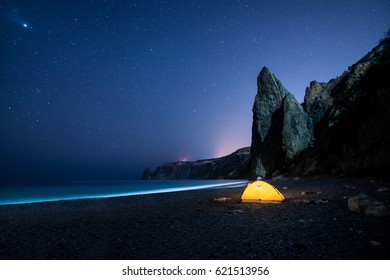 Glowing Camping Tent On A Beautiful Sea Shore With Rocks At Night Under A Starry Sky