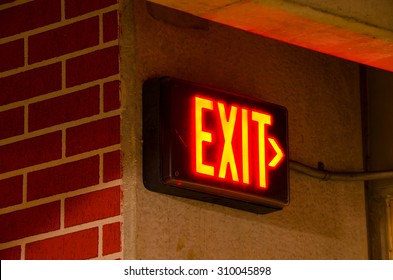 Glowing electrical exit sign mounted on a brick concrete wall at night in a dark corner of a corridor.