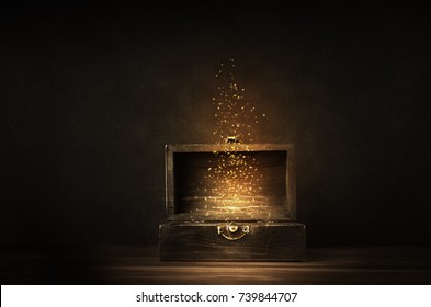 Glowing golden sparkles and stars rising from an old, opened wooden treasure chest. Darkly lit on a planked surface with black chalkboard background.