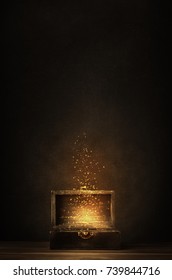 Glowing golden sparkles and stars rising from an old, opened wooden treasure chest. Darkly lit on a planked surface with black chalkboard background.