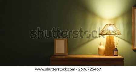 Glowing lamp with aroma reed diffuser, vase and frame on commode near green wall in dark room. Banner for design