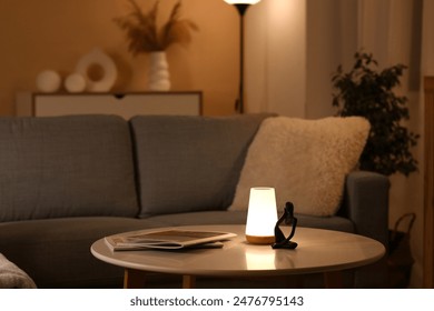 Glowing lamp on coffee table, comfortable sofa and magazines in stylish living room