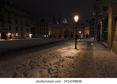 a glowing lantern from street lights on a snowy square and in the background is Prague Castle and St. Vitus Cathedral at night in Prague