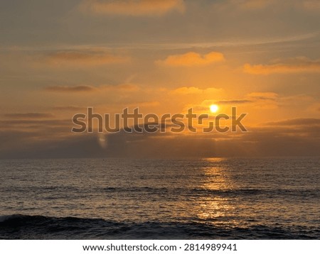 A glowing orange sun setting through a dramatic layer of stratified clouds above the dark waters of the Pacific Ocean in La Jolla, California.