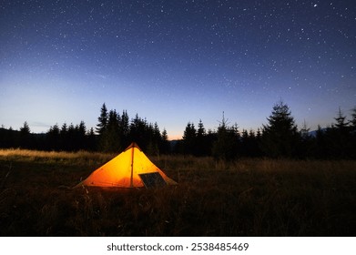 Glowing orange tent illuminated at dusk beside solar panel in grassy field. Backdrop of silhouetted trees and starry night sky emphasizes eco-friendly and serene camping experience. - Powered by Shutterstock