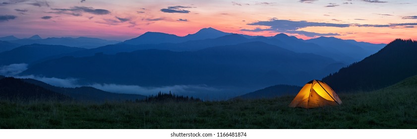 Glowing orange tent in the mountains under dramatic evening sky. Red sunset and mountains in the background. Summer landscape. Panorama