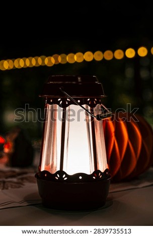Glowing red metal lantern with string lights in the background creates a warm ambiance, illustrating a seasonal centerpiece for festive evenings and enhancing the celebratory autumn atmosphere in a cozy outdoor setting. 