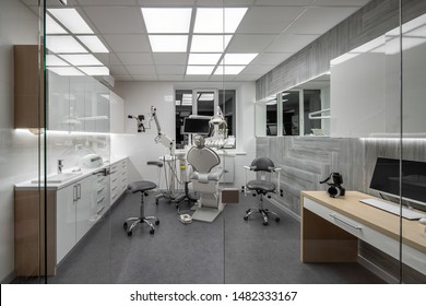 Glowing room in a dental clinic with light walls and dark gray floor. There is a workplace with a dental chair and equipment, table with a computer, zone with a sink, mirror, window, glass partition. - Shutterstock ID 1482333167