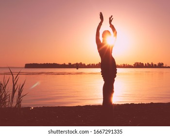 glowing silhouette of slim woman with hands up in the air illuminated with sunshine in front sky near river at sunrise