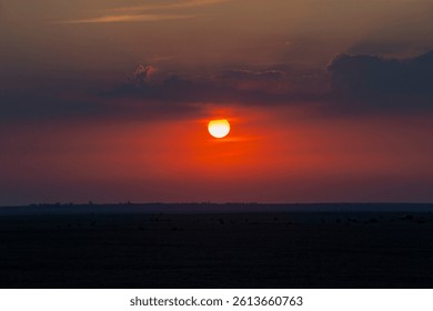 A glowing sun sets behind clouds, casting an orange hue over a vast, shadowy desert landscape with distant silhouettes.
