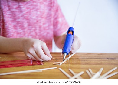 Glue Gun In The Child's Hand. Kid Glues Wooden Sticks With A Glue Gun. Closeup