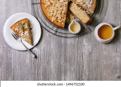 Gluten free almond rosemary homemade cake decorated by almond and sugar powder on cooling rack, with cup of tea, syrup and fork on over grey wooden background. Flat lay, copy space