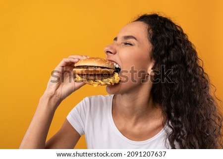 Gluttony. Closeup Portrait Of Funny Hungry Lady Biting Burger Eating Junk Food Posing Over Yellow Orange Studio Background. Woman Enjoying Big Hamburger. Unhealthy Nutrition And Bad Eating Habit