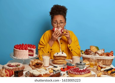 Gluttony And Overeating Concept. Upset Crying Ethnic Woman Eats Piece Of Cake Reluctantly, Sits At Table With Many Desserts, Isolated Over Blue Wall, Feels Hungry And Greedy, Wears Yellow Jacket