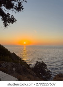A gnarled tamarix tree frames the sun over the Aegean Sea on Ikaria Island, Greece. Warm coastal twilight.
