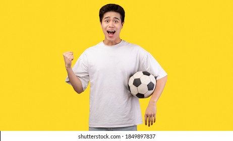 Go On. Portrait Of Excited Young Asian Man Making Winner Gesture, Watching Football Game, Standing And Holding Soccer Ball, Isolated Over Yellow Studio Background, Banner. Guy Cheering And Screaming