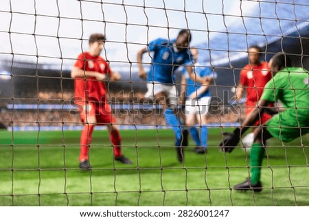 Goal net frames a motion-blurred attacker striking toward a waiting goalkeeper, capturing a scoring attempt as the goalie guards the post and tries to block the football attack