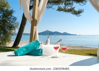 Goblets with rose champagne and rolled beach towels served on canopy bed prepared for romantic honeymoon on picturesque seashore in summer day