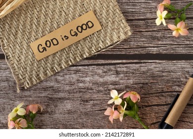 God Is Good, A Handwritten Text Quote On Hemp Textured Fabric With Flowers On Wooden Background In Vintage Style. The Biblical Concept Of The Love, Mercy, And Grace Of Jesus Christ. Top View.