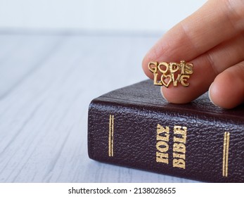 God Is Love, Text Quote With Golden Letter In Human's Hand Over Closed Holy Bible Book On A Gray Background With Copy Space. A Closeup. The Biblical Concept Of Grace And Mercy Of Jesus Christ.